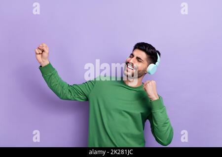 Foto di buon umore ragazzo allegro verde cuffie a manica lunga canto artista preferito danza pugni isolato su sfondo di colore viola Foto Stock