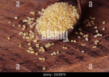 Asciugare Bulgur di grano in cucchiaio di legno rustico sfondo. Eventuali fuoriuscite di bulgur Foto Stock