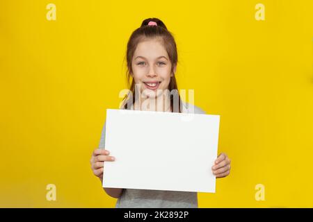 Ragazza carina con foglio bianco di carta su uno sfondo giallo. Bambina che tiene un foglio vuoto di carta Foto Stock