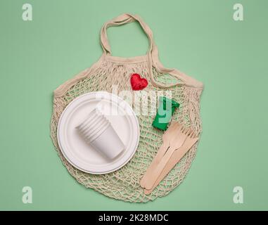 borsa di cotone bianco, tazze di carta e forchette e cucchiai di legno su sfondo verde. Rifiuti riciclabili, vista dall'alto Foto Stock