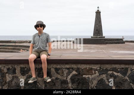 Ritratto di un adolescente in un cappello e occhiali da sole di fronte ad un piccolo faro. Bajamar. Tenerife. Isole Canarie. Spagna. Foto Stock