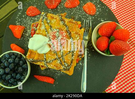 Cialde belghe tradizionali con mirtilli e fragole. Cioccolato fuso e panna montata sui waffle. Vista dall'alto. Sfondo nero in ardesia. Foto Stock