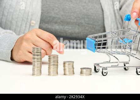 la donna siede al tavolo e tiene un carrello in miniatura e pile di monete bianche. Concetto di risparmio, vendita, imposta Foto Stock