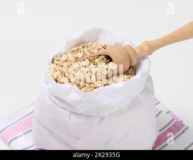 farinata d'avena cruda in un sacchetto di carta bianca e un cucchiaio di legno su un tavolo bianco, porridge per la colazione Foto Stock