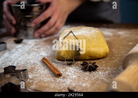 Un taglialegna a forma di albero di Natale è appoggiato contro l'impasto, una foto in chiave scura, altre muffe sono visibili, una spilla, un setaccio di ferro per la farina, che è tenuto nelle mani di un pasticciere. Foto Stock