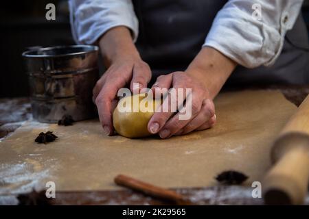 Sulla pergamena è presente un impasto leggermente cosparso di farina, che il cuoco tiene in mano, un matterello, taglierine per biscotti, cannella e un vaso di farina metallica sono visibili intorno. Foto Stock