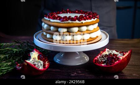 Una torta a tre strati, composta da torte e crema bianca, decorata con melograno e rosmarino in cima, si erge su un supporto bianco lateralmente su uno sfondo scuro Foto Stock