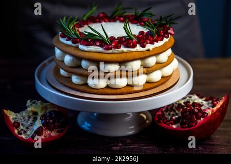 Una torta a tre strati, composta da torte e crema bianca, decorata con melograno e rosmarino in cima, si erge su un supporto bianco lateralmente su uno sfondo scuro Foto Stock