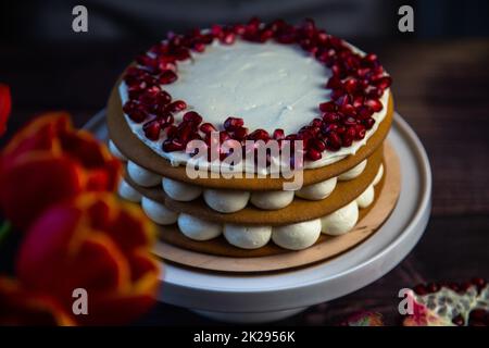 Su fondo scuro, accanto a tulipani e melograni, sorge una torta a tre strati composta da torte e crema bianca, decorata con melograno in cima a cerchio. Foto Stock