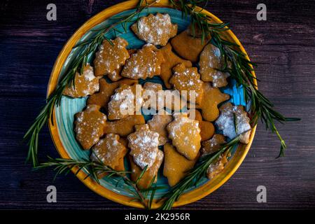I biscotti sotto forma di alberi di Natale giacciono su un piatto blu, decorato con rosmarino ai lati su uno sfondo scuro Foto Stock