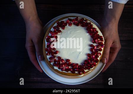 Una torta a tre strati, composta da torte e crema bianca, decorata con melograno sulla sommità, è tenuta nelle mani di un cuoco su uno sfondo scuro, vista dall'alto Foto Stock