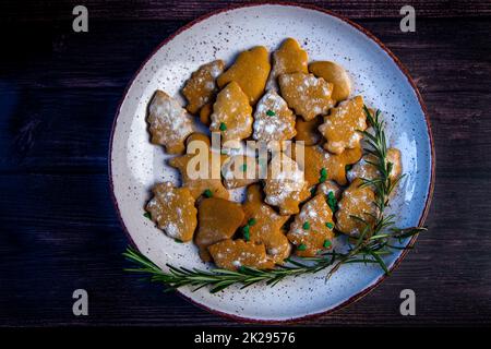 I biscotti sono su un piatto di forme diverse, in primo piano c'è un biscotto a forma di albero di Natale, decorato con zucchero a velo come neve e piccoli alberi verdi di Natale sullo sfondo del resto dei biscotti, un ramo di rosmarino Foto Stock