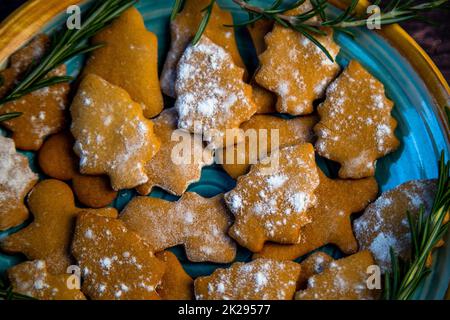 I biscotti sotto forma di alberi di Natale giacciono su un piatto blu, decorato con rosmarino ai lati su uno sfondo scuro Foto Stock
