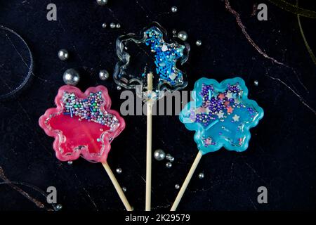 I lollipop su un bastone di legno a forma di sonaglino trasparente, rosa e blu si trovano su uno sfondo di marmo scuro circondato da nastri d'oro e d'argento e palle d'argento Foto Stock