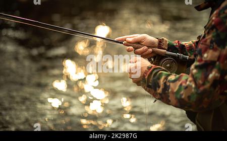 Vista ravvicinata delle mani di un pescatore a mosca che lavorano la linea e la canna da pesca mentre la pesca con la mosca su di uno splendido fiume di montagna per la trota iridea Foto Stock