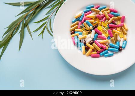 Piatto bianco con pillole di integratori alimentari in diversi colori luminosi. Sfondo blu, posto per un'iscrizione. Ramo verde sullo sfondo. Foto Stock