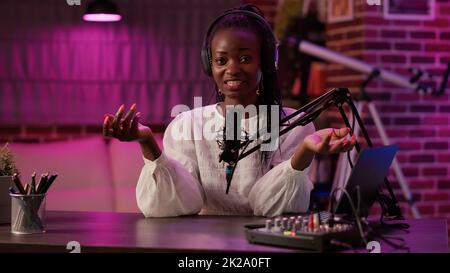 Donna afro-americana vlogger parlare con il pubblico mentre registrare podcast utilizzando mixer audio e microfono. Famoso influencer che crea contenuti per la pagina dei social media online da podcasting studio. Foto Stock