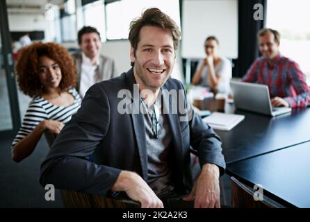 Abbiamo stabilito il livello del successo. Ritratto di un lavoratore in ufficio seduto a un tavolo della sala riunioni con i colleghi in background. Foto Stock