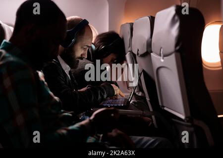 Gruppo multietnico di persone che volano in classe economica insieme per arrivare a destinazione, uomo che utilizza il computer portatile durante il volo internazionale. Turisti che viaggiano all'estero in aereo. Foto Stock