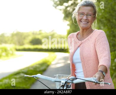 Gode di attività all'aperto sane - Ciclismo. Sorridente donna anziana in piedi su una corsia di campagna con la sua bicicletta. Foto Stock