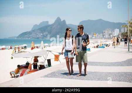 Godendo la spiaggia con un freind. Scatto corto di una giovane coppia che cammina lungo una spiaggia trafficata. Foto Stock