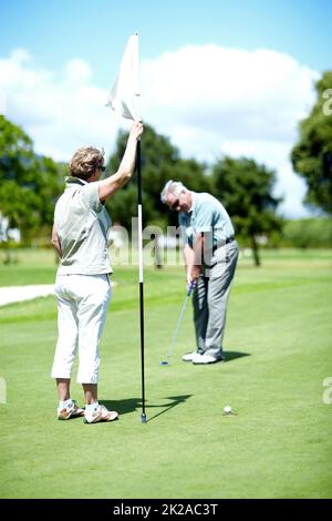 Master del foro in uno. Una donna anziana che detiene una bandiera mentre suo marito mette per il birdie. Foto Stock