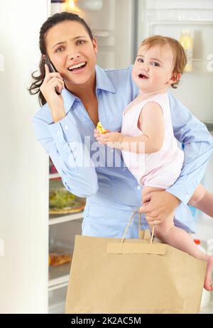 Troppo a volte per destreggiarsi. Una mamma che lavora sopraffatta che parla al telefono mentre tiene il suo bambino e una borsa di generi alimentari. Foto Stock