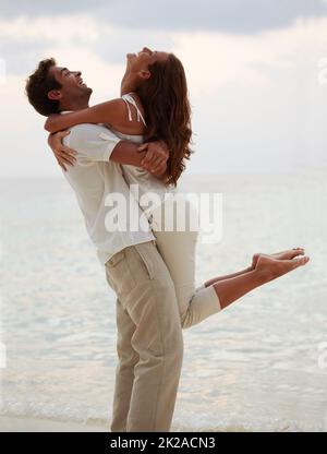 Un abbraccio serale - Romance. Giovane che solleva la sua splendida ragazza in aria mentre si trova sulla spiaggia. Foto Stock