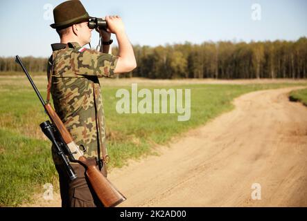 Alla vista. Un ranger di gioco con il suo fucile guardando attraverso il suo binocolo all'aperto con copyspace. Foto Stock