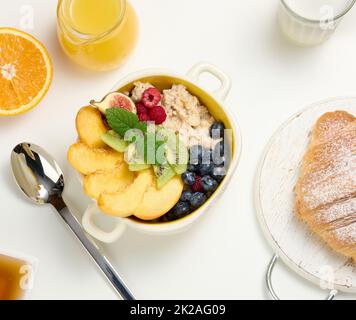 piatto con farina d'avena e frutta, mezzo succo d'arancia maturo e spremuto di fresco in un decanter trasparente in vetro, miele in una ciotola su un tavolo bianco. Foto Stock