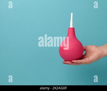 una mano in un guanto medico blu tiene un enema di gomma rosa su uno sfondo blu Foto Stock