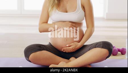 In buone mani. Scatto corto di una giovane donna incinta irriconoscibile strofinando il suo bump del bambino mentre si siede su un tappeto di yoga. Foto Stock