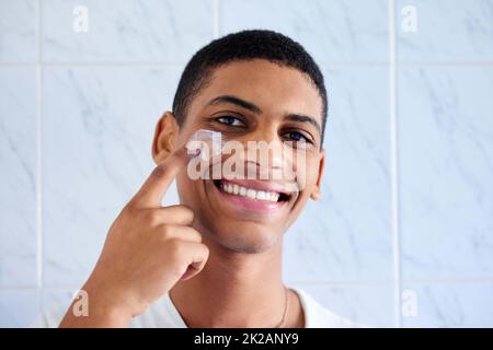 Anche la cura della pelle per uomo è importante. Ritratto corto di un bel giovane uomo in piedi da solo nel suo bagno e applicare crema viso. Foto Stock