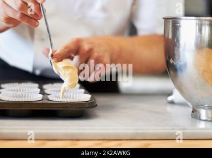 Pastella su. Un primo piano di un panettiere che spinge l'impasto per cupcake da un cucchiaio nel vassoio per cupcake Foto Stock