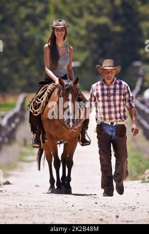 Equitazione 101. Una giovane cowgirl attraente che guida un cavallo su un ranch mentre un cowboy cammina accanto. Foto Stock