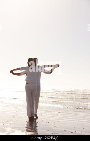 Rimboccati alla luce del mattino. Scatto a tutta lunghezza di una giovane coppia attraente vestita di bianco camminando lungo una spiaggia. Foto Stock