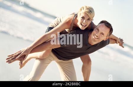 Per sempre divertente. Un marito che dà a sua moglie una cavalcata sulla spiaggia. Foto Stock