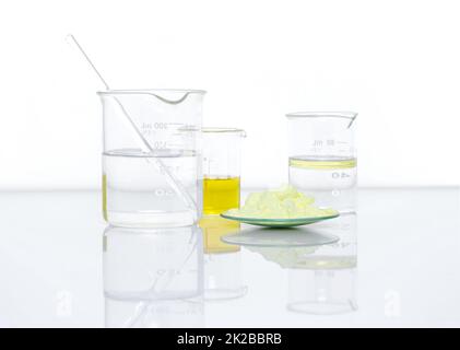 Ingrediente chimico di primo piano su tabella bianca del laboratorio. Polvere di zolfo in vetro chimico da orologio posizionare accanto al cloruro di alluminio liquido, olio e alcool in becher. Vista laterale Foto Stock