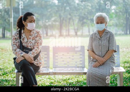 Donna anziana asiatica e figlia in sociale distanziamento seduta panca e indossare maschera viso per proteggere l'infezione di sicurezza Covid-19 Coronavirus in parco. Foto Stock