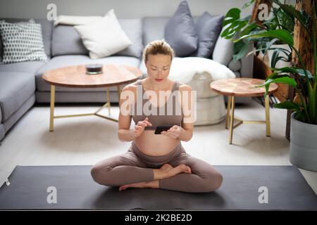 Gravidanza e tecnoloy supportato sano stile di vita concetto. Allegro felice donna incinta utilizzando l'applicazione smartphone mentre si esercita sul tappetino yoga sul pavimento del soggiorno a casa. Foto Stock