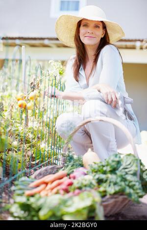 Cena gratuita con cortesia della terra. Una bella donna si accoccola nel suo orto con un cesto di verdure appena raccolte davanti a lei. Foto Stock