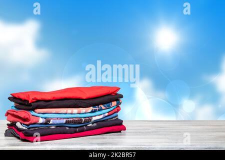 Impilare abiti colorati. Primo piano di un mucchio di t-shirt o camicie colorate su un tavolo luminoso contro astratto sfondo del cielo blu sfocato e soleggiato. Spazio per la registrazione del display del prodotto. Moda estiva. Foto Stock