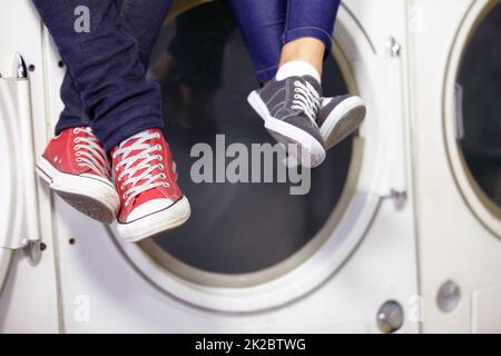 Si rilassò mentre si faceva il bucato. Immagine ritagliata di una coppia piedi come si siedono su una lavatrice alla lavanderia automatica. Foto Stock