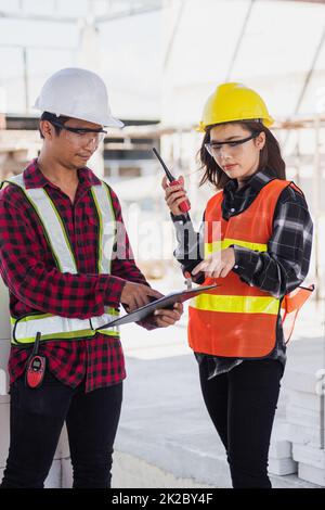 Due ingegneri asiatici capimana architetto operaio uomo e donna che parlano in cantiere, ingegneria tenere discussione radio gestire progetto e controllare lavoratore dipendente di costruzione Foto Stock