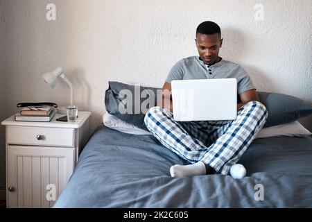 Quale serie dovrei guardare più tardi. Scatto a tutta lunghezza di un bel giovane uomo seduto da solo sul suo letto e utilizzando il suo computer portatile a casa. Foto Stock