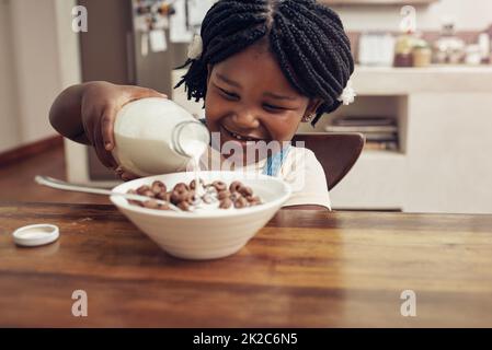 Mi piace la colazione molto lattiginosa. Il colpo corto di una ragazza piccola adorabile che versa il latte nella sua ciotola del cereale a casa. Foto Stock