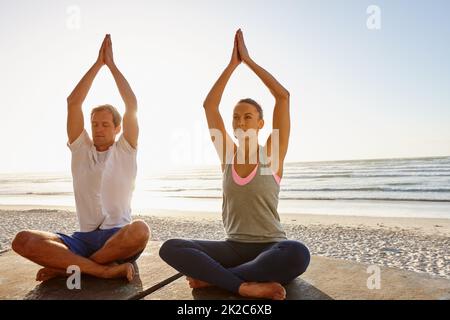 Il theyre che conduce uno stile di vita equilibrato. Scatto di una coppia che fa yoga in spiaggia al tramonto. Foto Stock