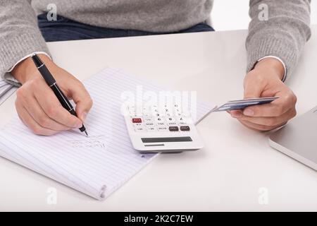 Calcolo di ciò che ho speso questo mese. Un primo piano di un uomo in possesso di una carta di credito che scrive qualcosa sul suo blocco note mentre usa una calcolatrice. Foto Stock