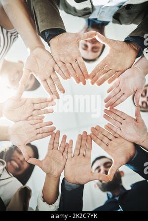 Insieme i loro cervelli funzionano come uno. Inquadratura ad angolo basso di un gruppo di uomini d'affari allegri che formano una huddle con le mani e che guardano verso il basso all'interno dell'ufficio. Foto Stock