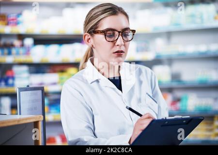 Prendere appunti di tutte le scorte mediche in magazzino. Shot di un giovane farmacista che scrive su una clipboard in una farmacia. Foto Stock